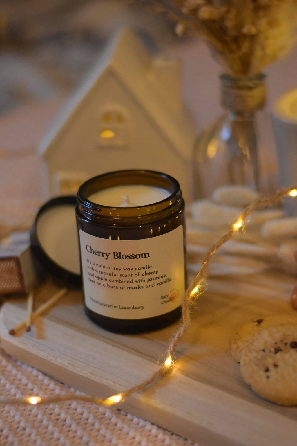 Bougie en cire de soja avec couvercle, posée sur une planche en bois, accompagnée de cookies, d’allumettes, d’une guirlande lumineuse et d’une décoration cosy.