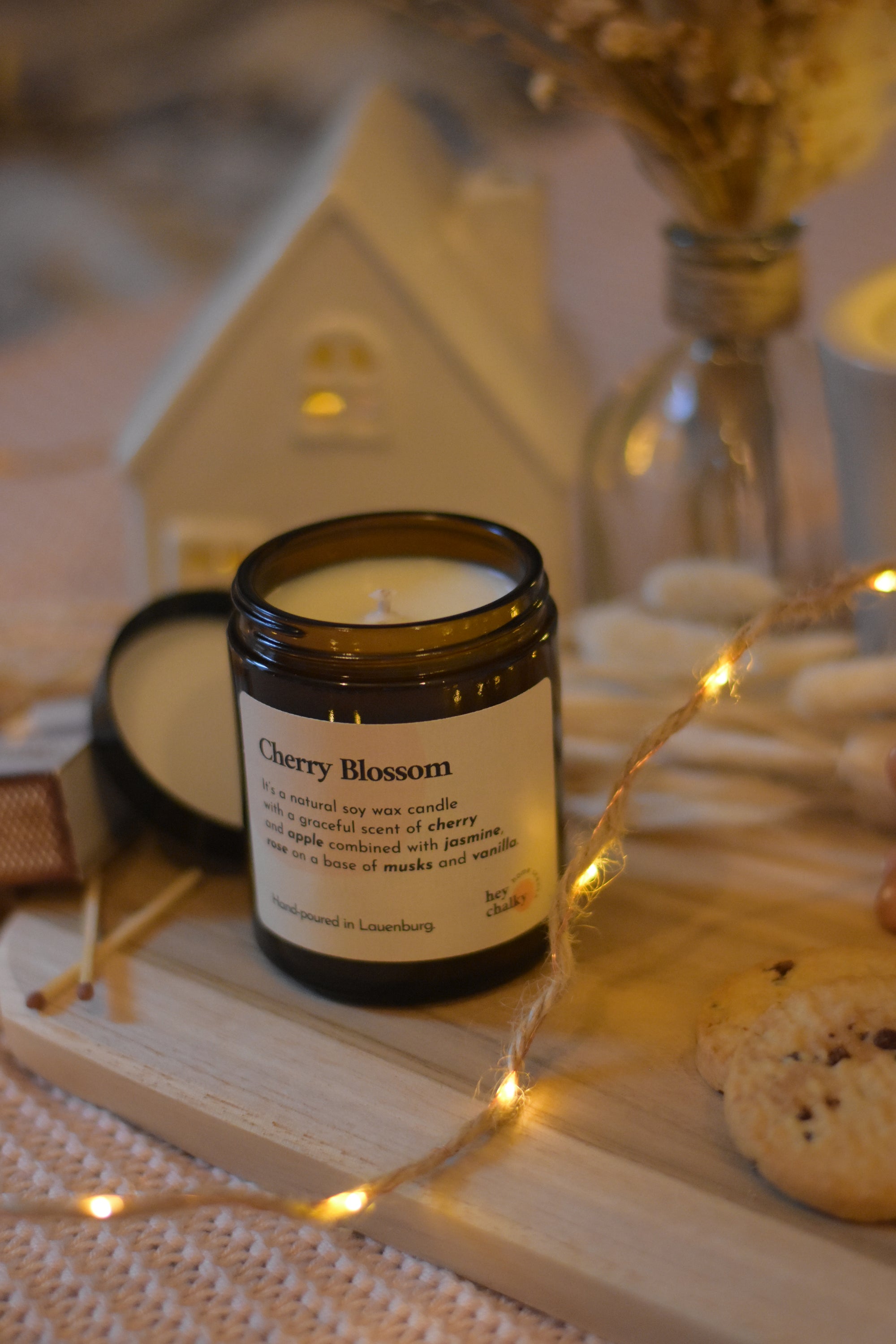 Bougie en cire de soja avec couvercle, posée sur une planche en bois, accompagnée de cookies, d’allumettes, d’une guirlande lumineuse et d’une décoration cosy.