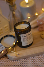 Bougie en cire de soja avec mèche en coton, fermée par un couvercle, posée sur une planche en bois et un plaid rose, avec des allumettes à côté et une guirlande lumineuse.