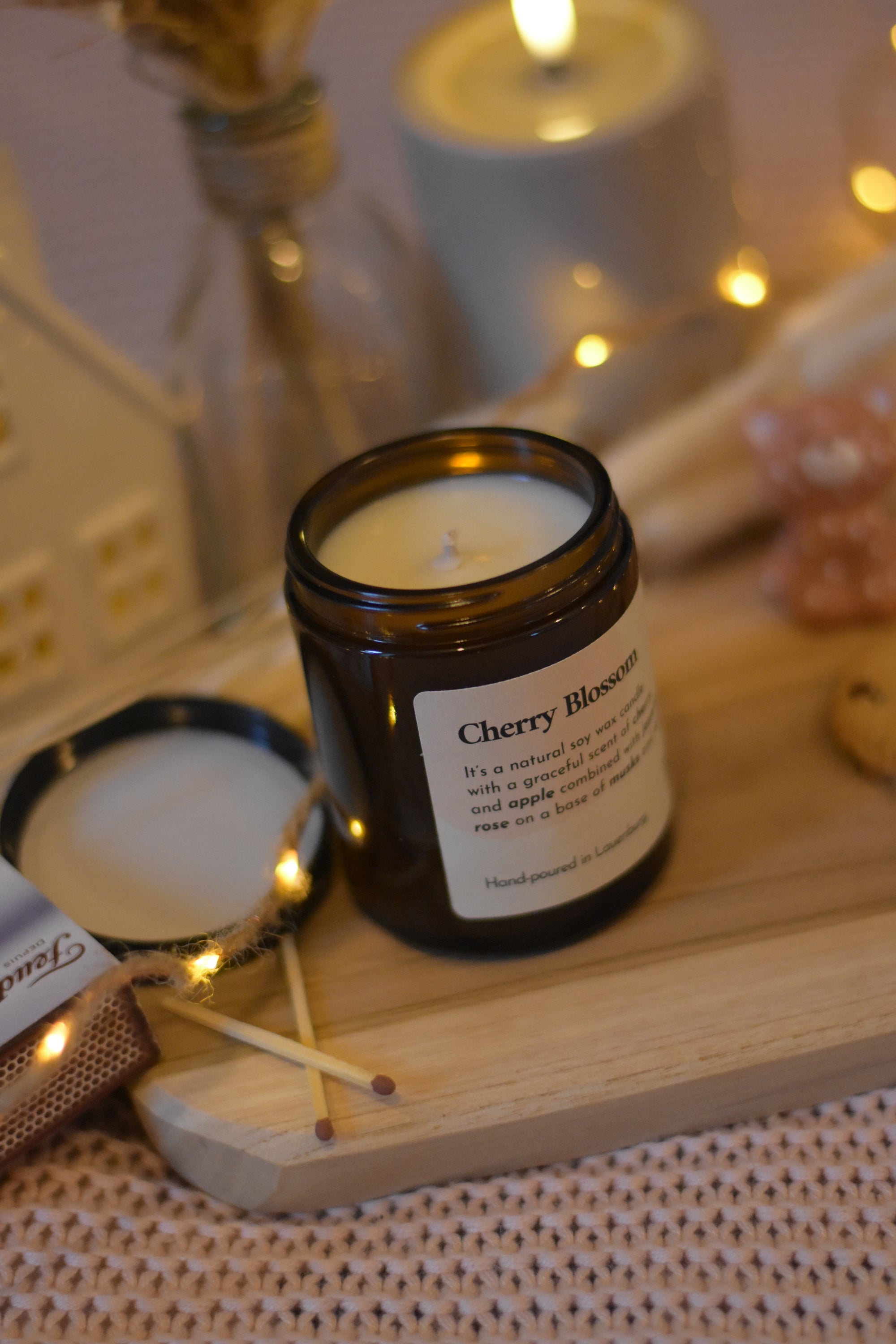 Bougie en cire de soja avec mèche en coton, fermée par un couvercle, posée sur une planche en bois et un plaid rose, avec des allumettes à côté et une guirlande lumineuse.