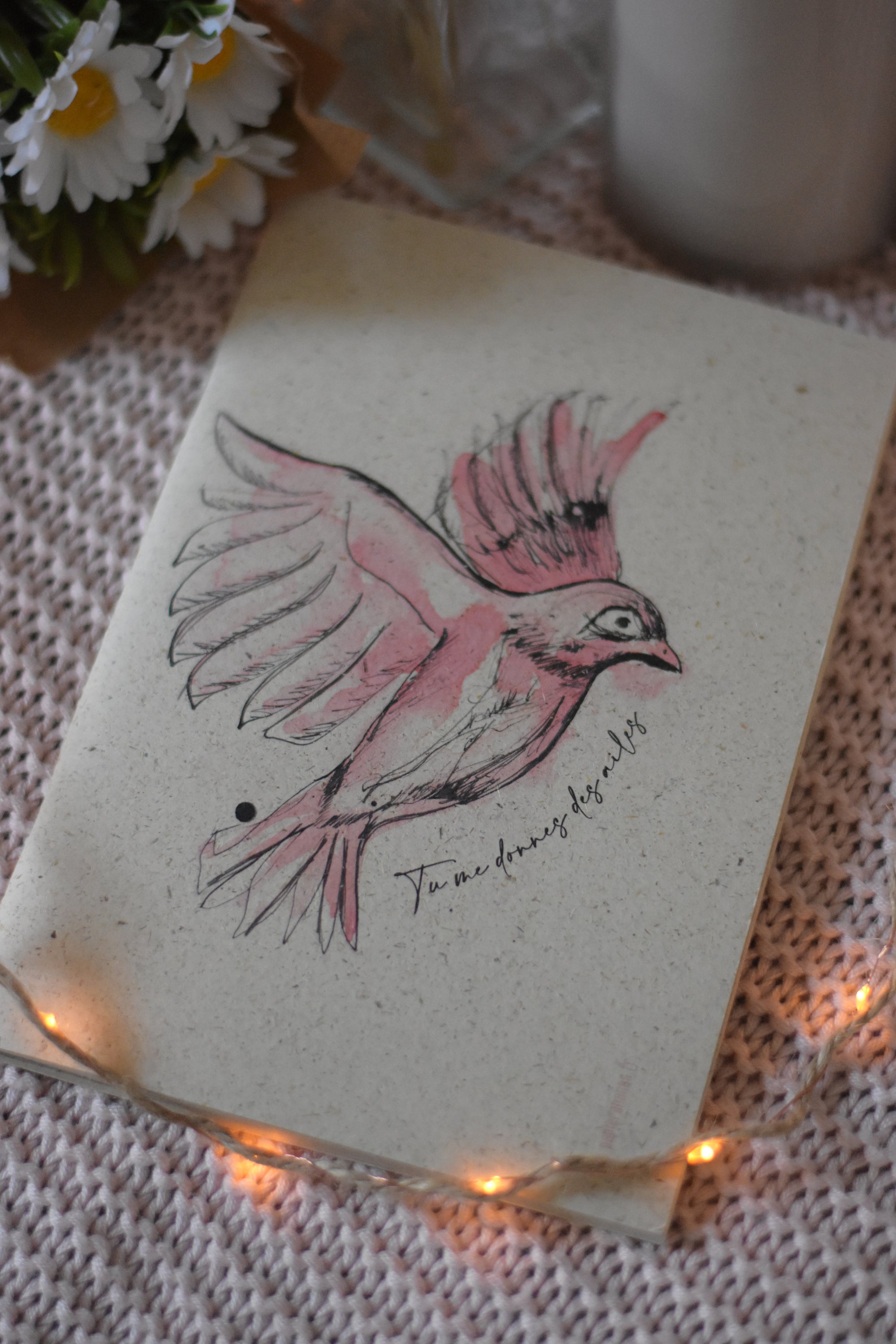Carnet de note en papier feuille illustrée d’un oiseau rose en plein vol, posée sur un plaid tricoté rose avec une guirlande lumineuse et un bouquet de marguerites en arrière-plan