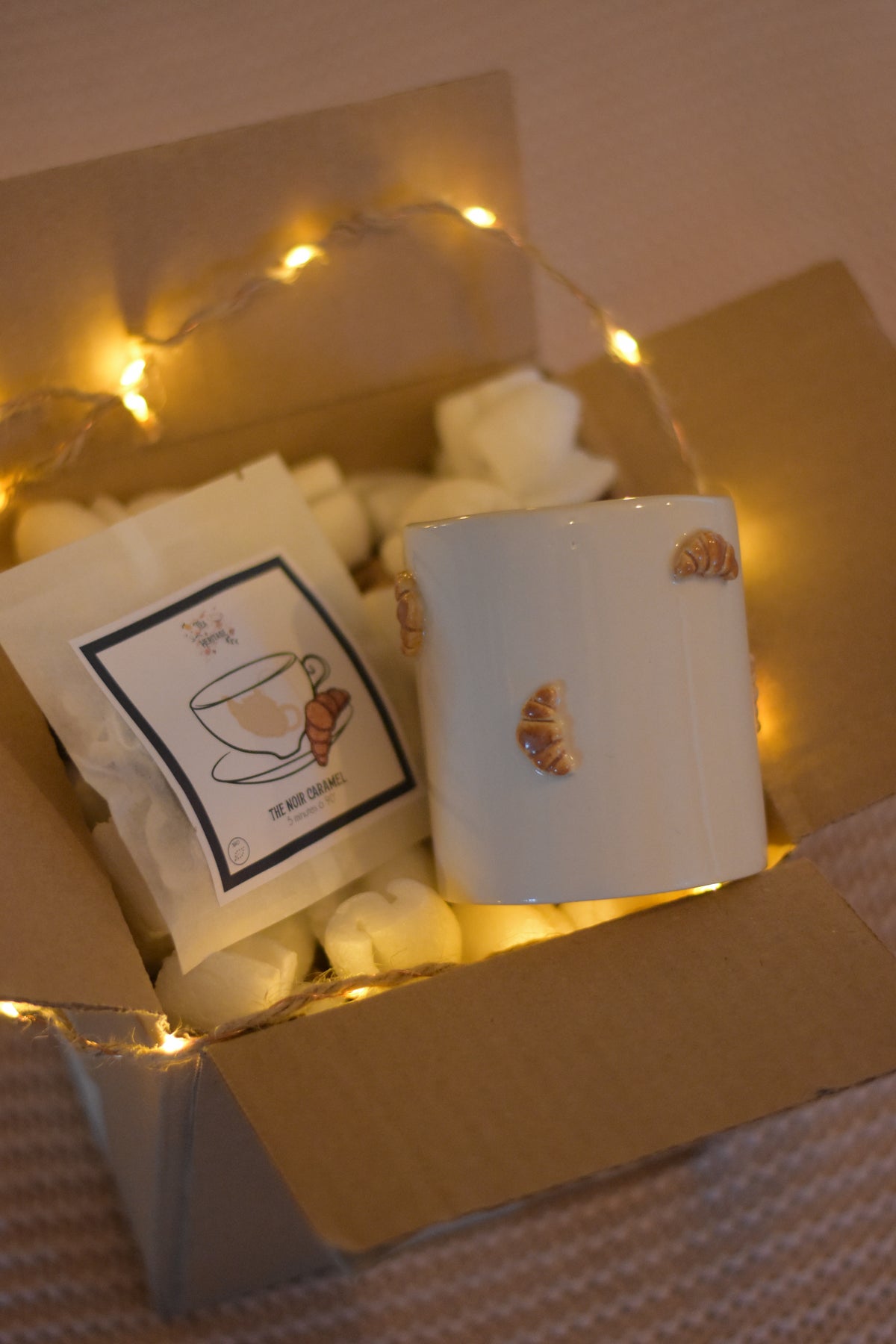 Coffret cosy posé sur un plaid rose. Il contient 5 sachets de thé bio au caramel et un mug artisanal fait à la main, décoré de petits croissants en relief, accompagné d’une guirlande lumineuse.