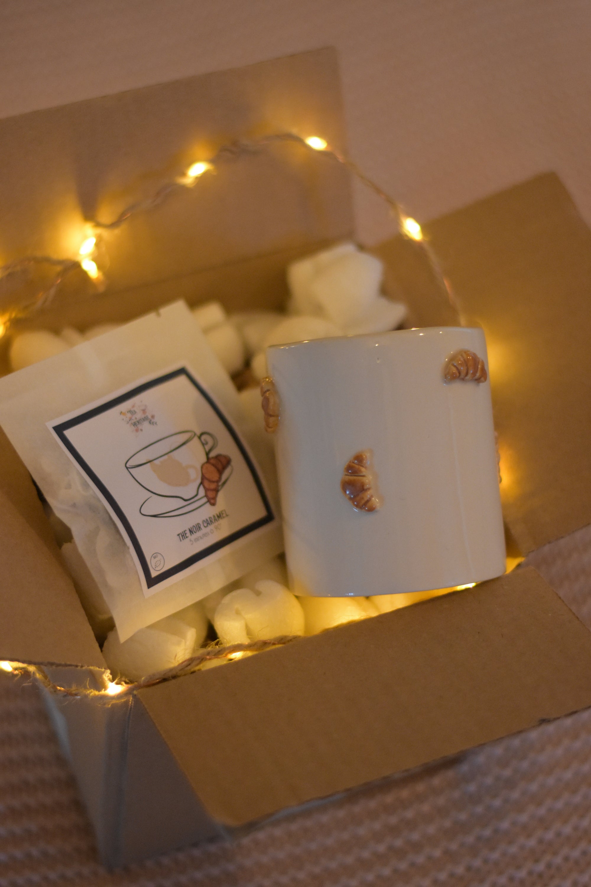 Coffret cosy posé sur un plaid rose. Il contient 5 sachets de thé bio au caramel et un mug artisanal fait à la main, décoré de petits croissants en relief, accompagné d’une guirlande lumineuse.