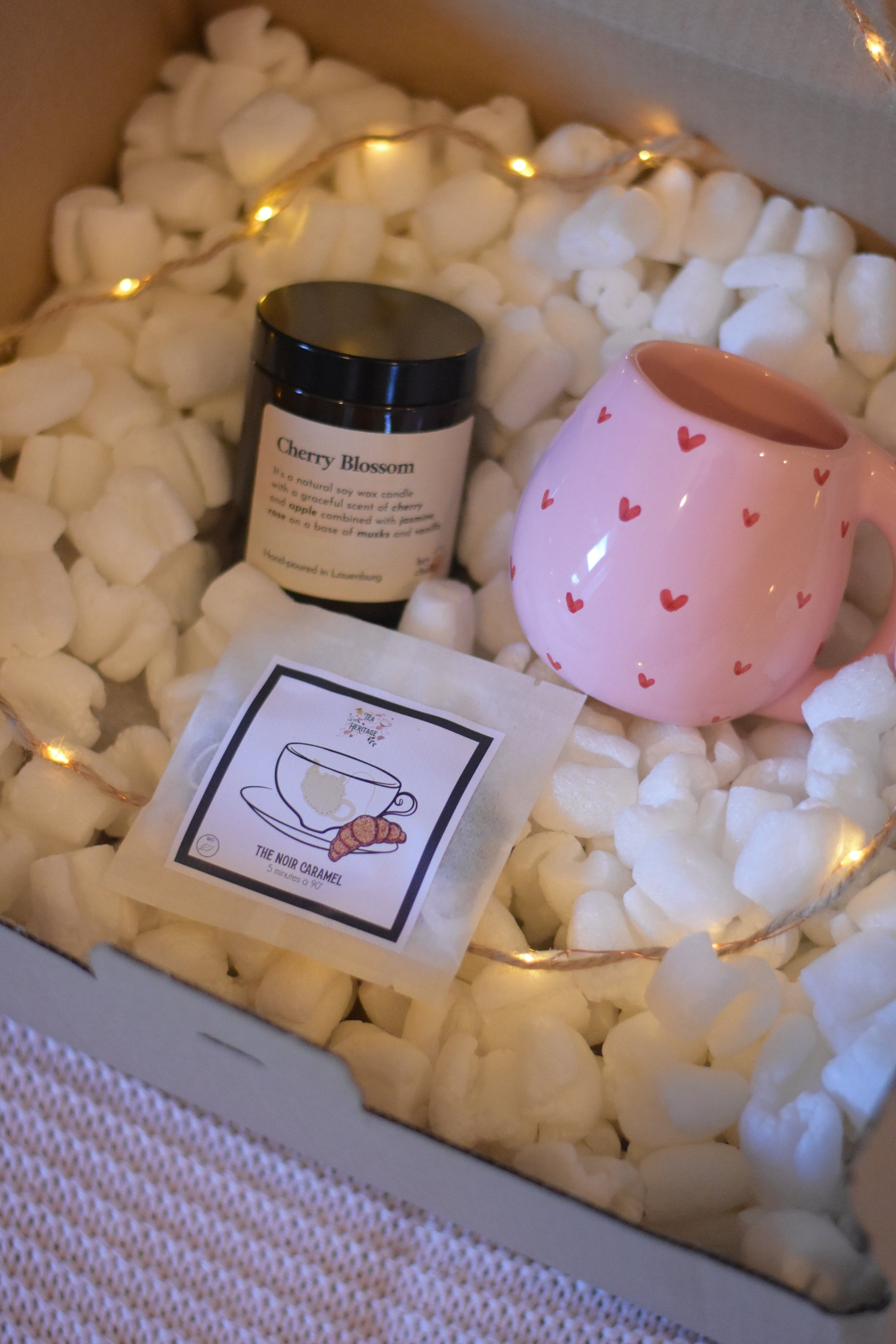 Coffret cosy contenant un mug artisanal, du thé bio au caramel et une bougie en cire de soja avec mèche en coton dans un pot en verre, le tout accompagné d’une guirlande lumineuse