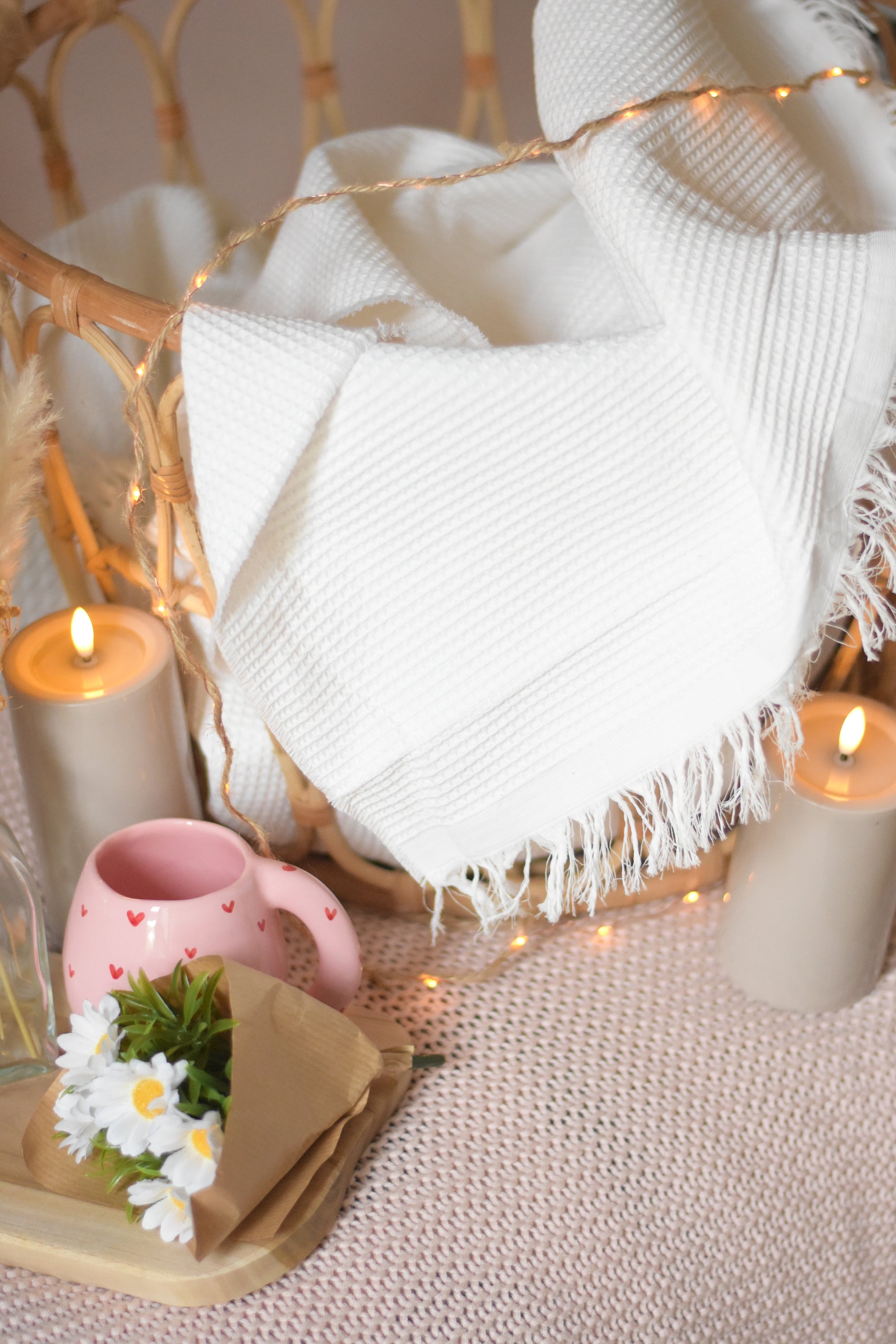 Plaid blanc à franges présenté dans un panier en rotin, accompagné d’un mug rose à cœurs, de bougies allumées et d’une guirlande lumineuse pour illustrer une ambiance chaleureuse et cosy.