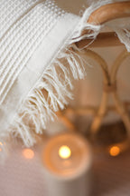 Plaid blanc en nid d’abeille avec franges, dans un panier en rotin, accompagné d’une bougie et d’une guirlande lumineuse.