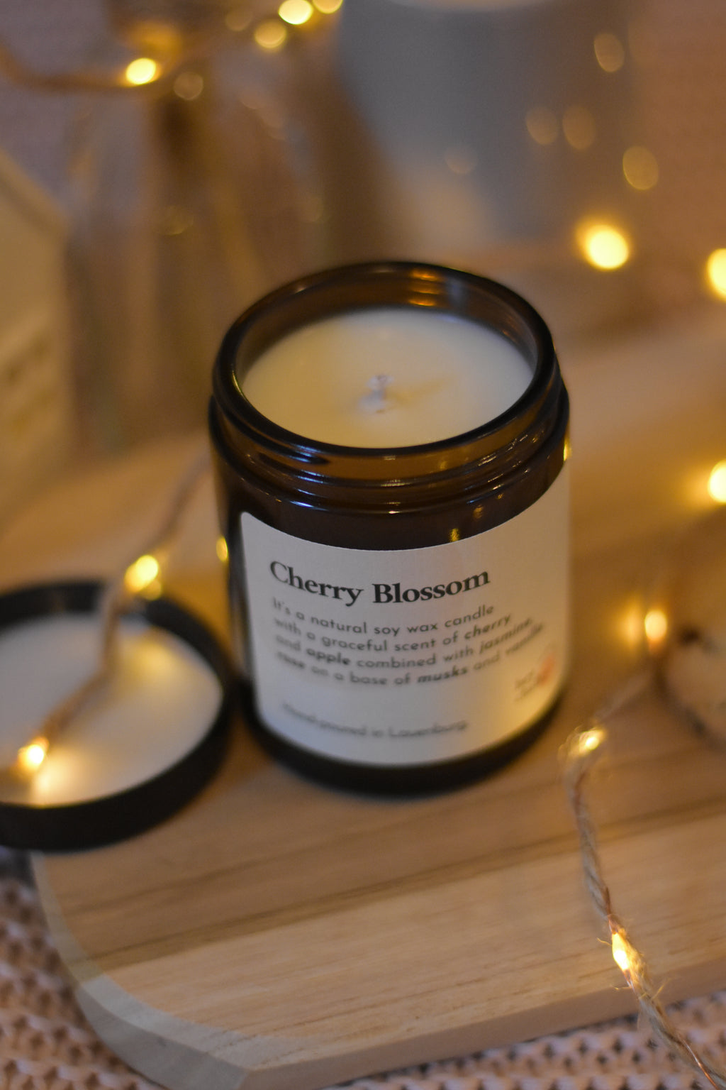 Bougie à la cire de soja avec mèche en coton dans un pot en verre ambré, posée sur une planche en bois, accompagnée de son couvercle, d’une guirlande lumineuse et d’une ambiance cosy.