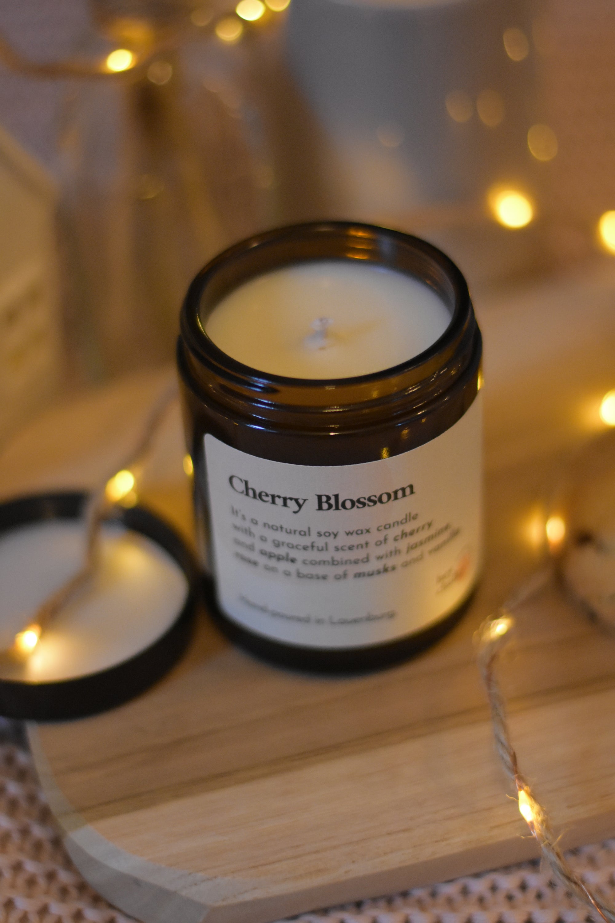 Bougie à la cire de soja avec mèche en coton dans un pot en verre ambré, posée sur une planche en bois, accompagnée de son couvercle, d’une guirlande lumineuse et d’une ambiance cosy.