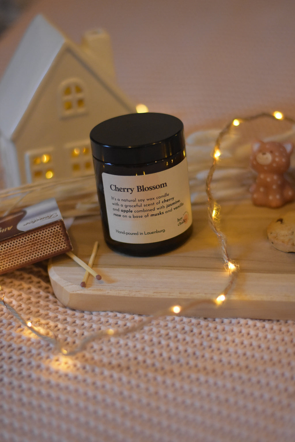 Bougie en cire de soja avec mèche en coton, fermée par un couvercle, posée sur une planche en bois et un plaid rose, avec des allumettes à côté et une guirlande lumineuse.