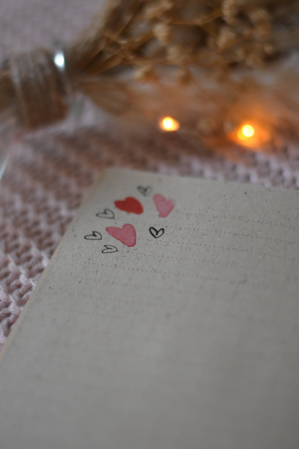 Carnet de notes ouvert posé sur un plaid rose avec une illustration de petit cœur rose et rouge en haut de page, posé à côté de fleurs séchées et d’une guirlande lumineuse.