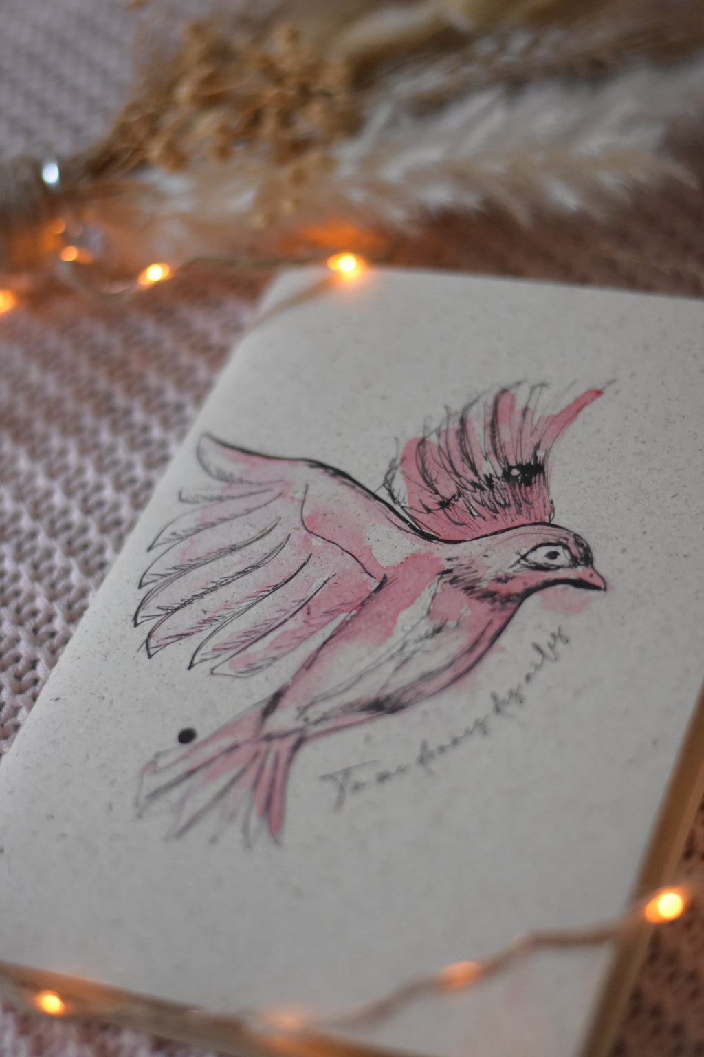 Carnet de notes posé sur un plaid rose, entouré d’une guirlande lumineuse et de fleurs séchées.