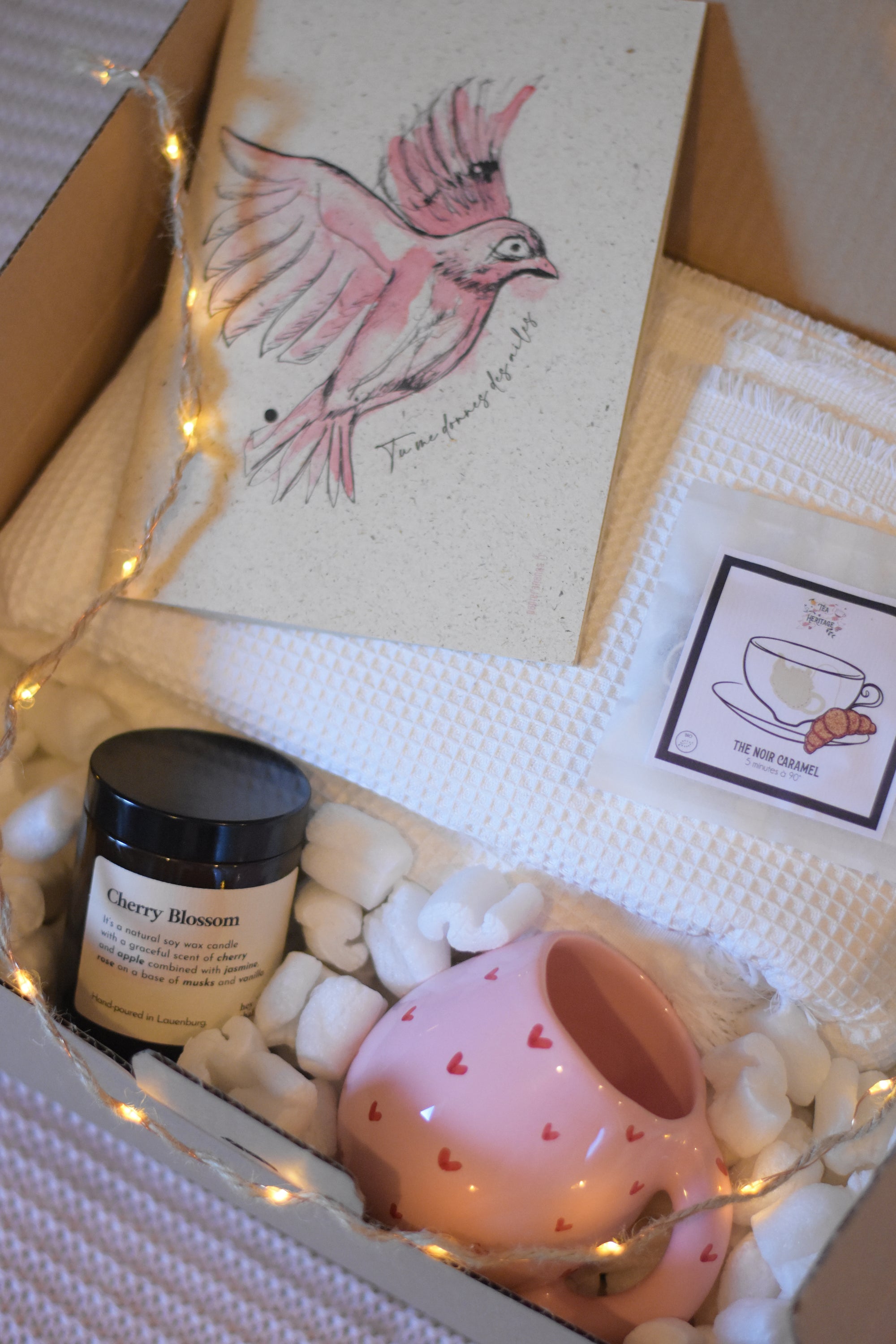 Coffret cosy posé sur un plaid rose, contenant un carnet de notes décoré d’un oiseau, un plaid blanc en nid d’abeille, une tasse ou mug rose avec de petits cœurs rouges, une bougie avec couvercle dans un pot en verre, du thé bio et une guirlande lumineuse.