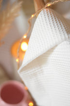 Plaid blanc en nid d’abeille dans un panier en rotin, accompagné d’une guirlande lumineuse, d'un mug rose et de fleurs séchées.