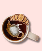 Sachet de thé en forme de théière avec un croissant pailleté, placé dans une tasse de thé chaud, le tout dans un décor rose.