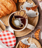 Sachet de thé en forme de théière décorée d’un petit croissant pailleté, présenté dans une tasse de thé chaude avec un croissant frais en arrière-plan, ambiance petit-déjeuner gourmand