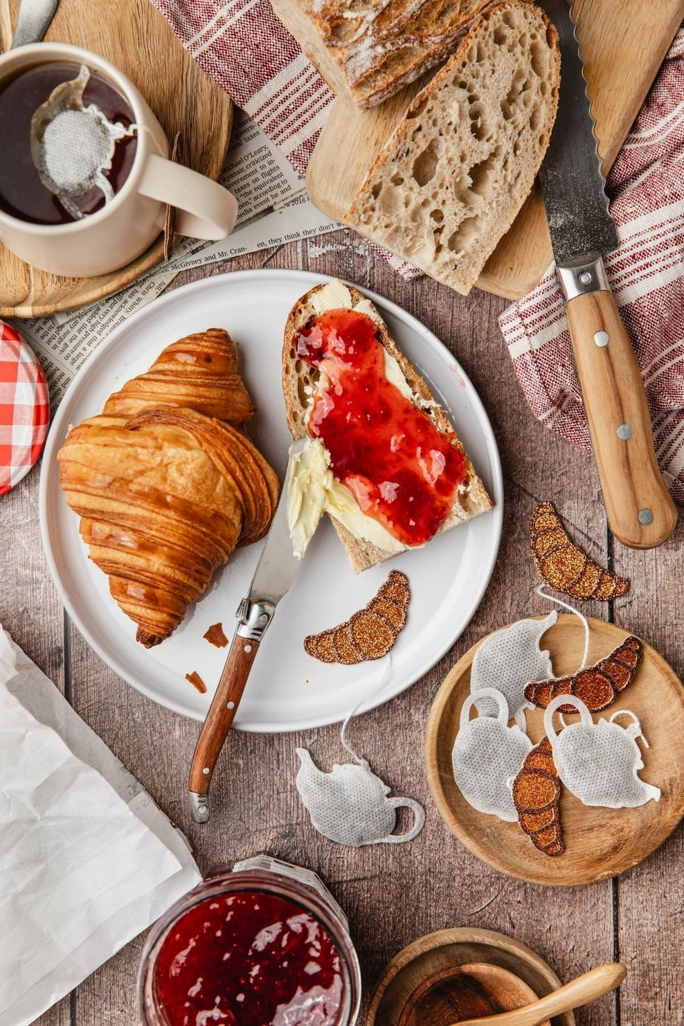 Sachet de thé en forme de théière et petit croissant pailleté, dans une ambiance de petit-déjeuner gourmand avec croissants, pain, confiture et tasse de thé.